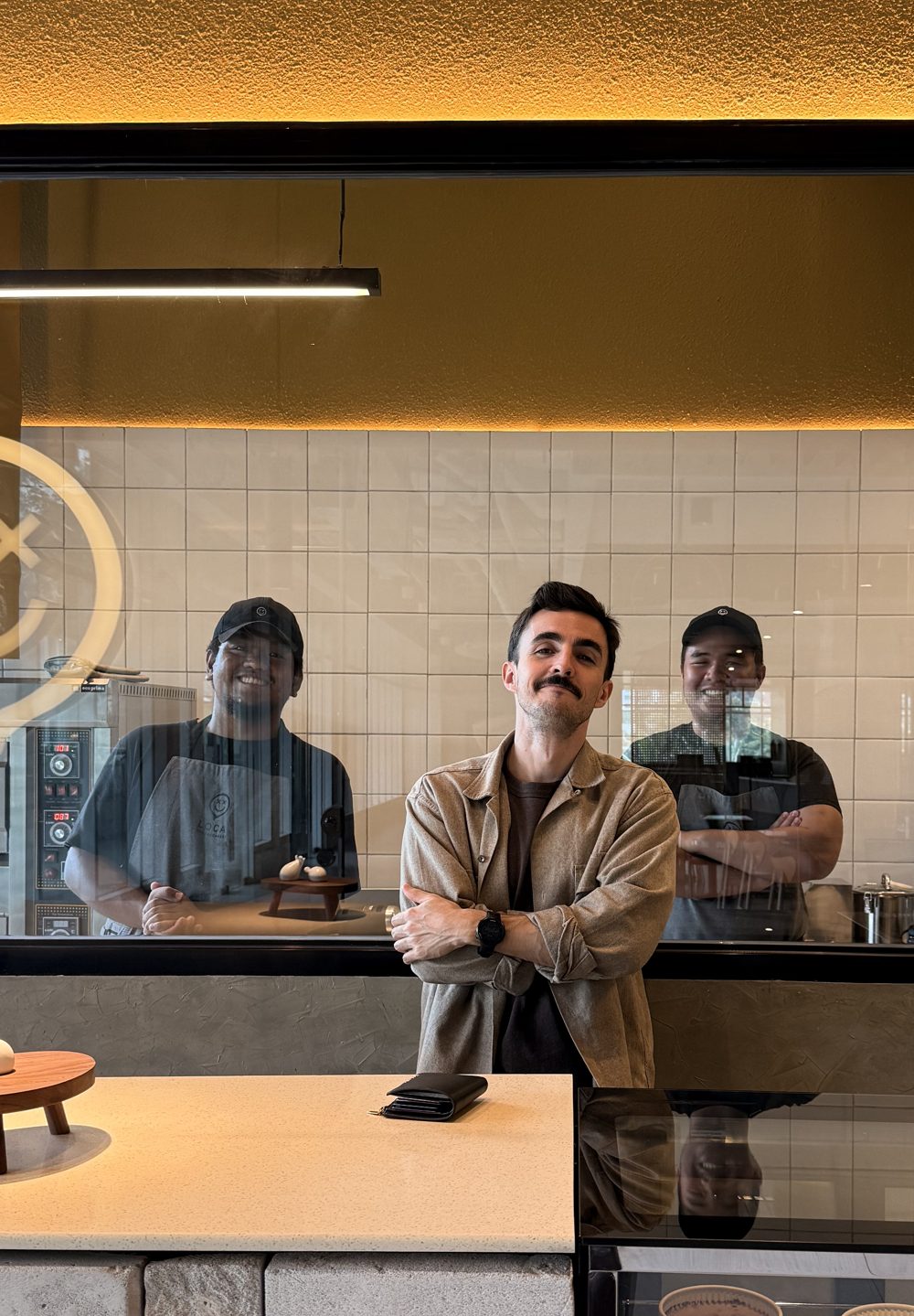 Gonzalo at the Loca Cheesecakes store with his two bakers smiling behind the kitchen window
