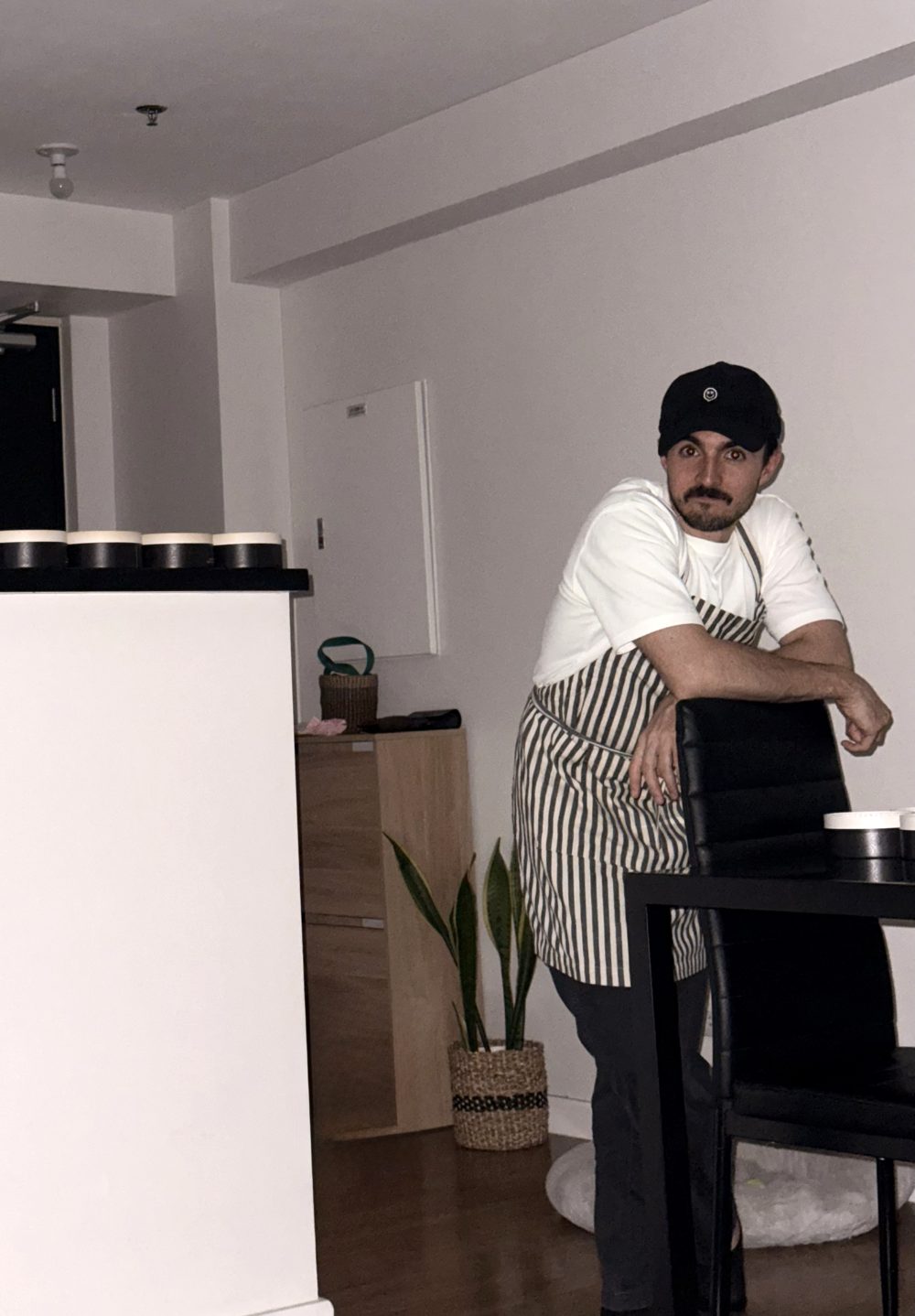 Gonzalo baking at home in an apron, leaning on a chair, with cheesecakes lined up on the counter