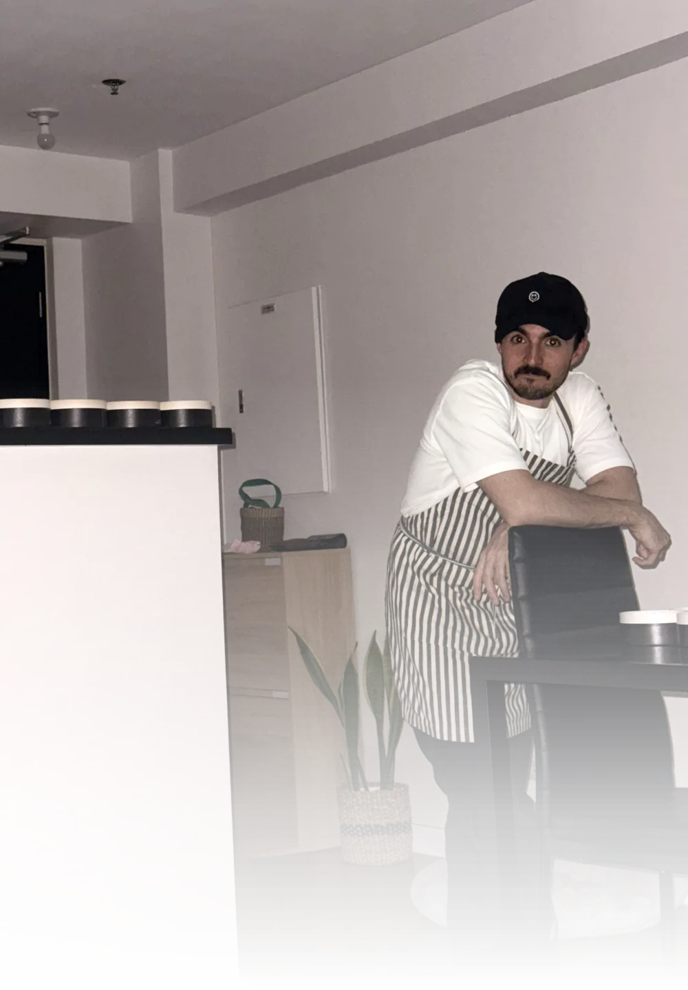 Gonzalo baking at home in an apron, leaning on a chair, with cheesecakes lined up on the counter